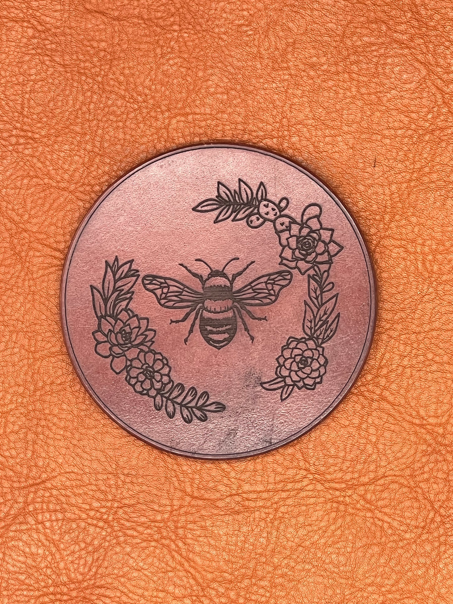 a chestnut colored leather coaster featuring an engraved bee flanked with flowers