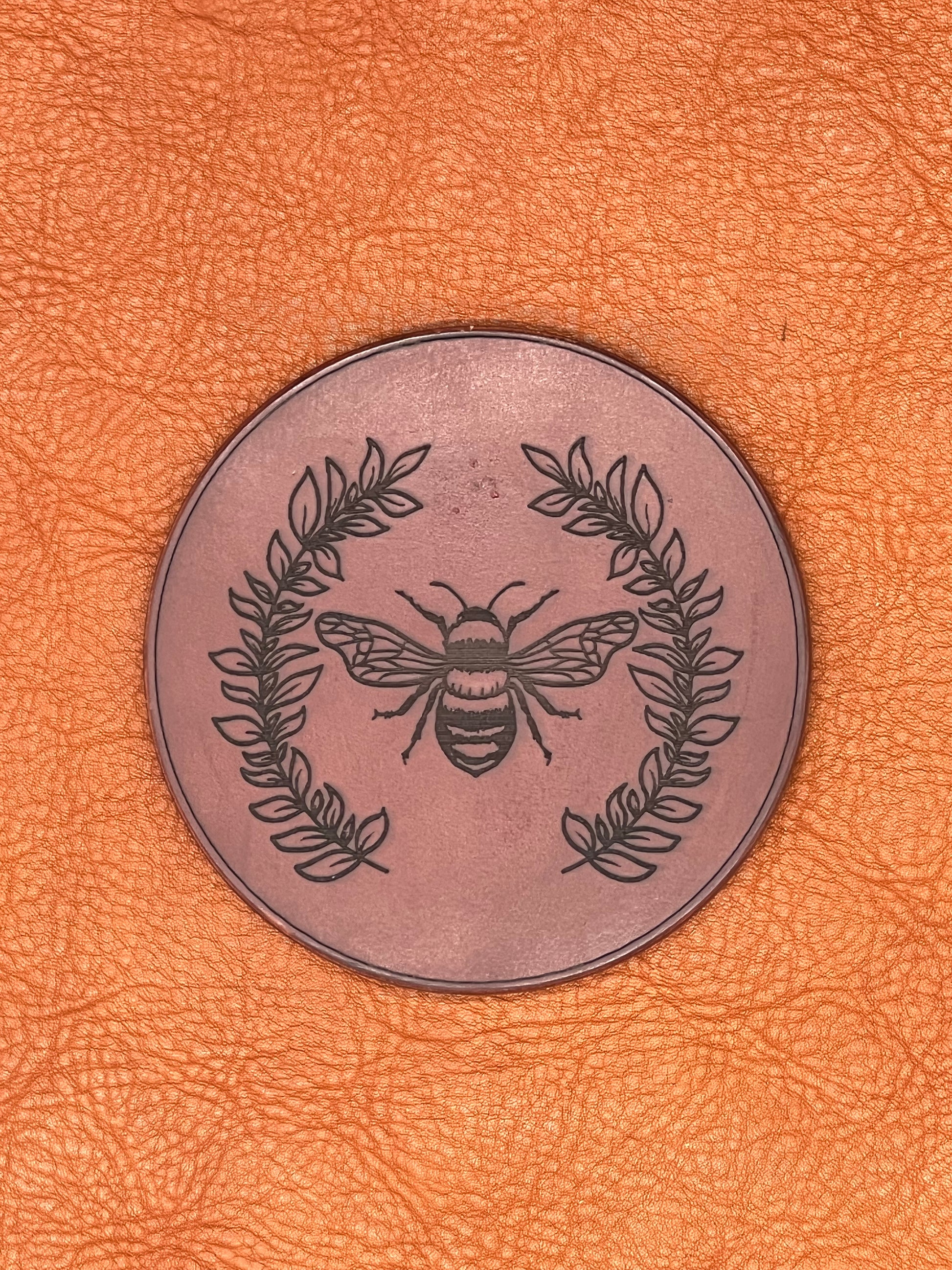 a chestnut colored leather coaster featuring an engraved bee flanked with vines