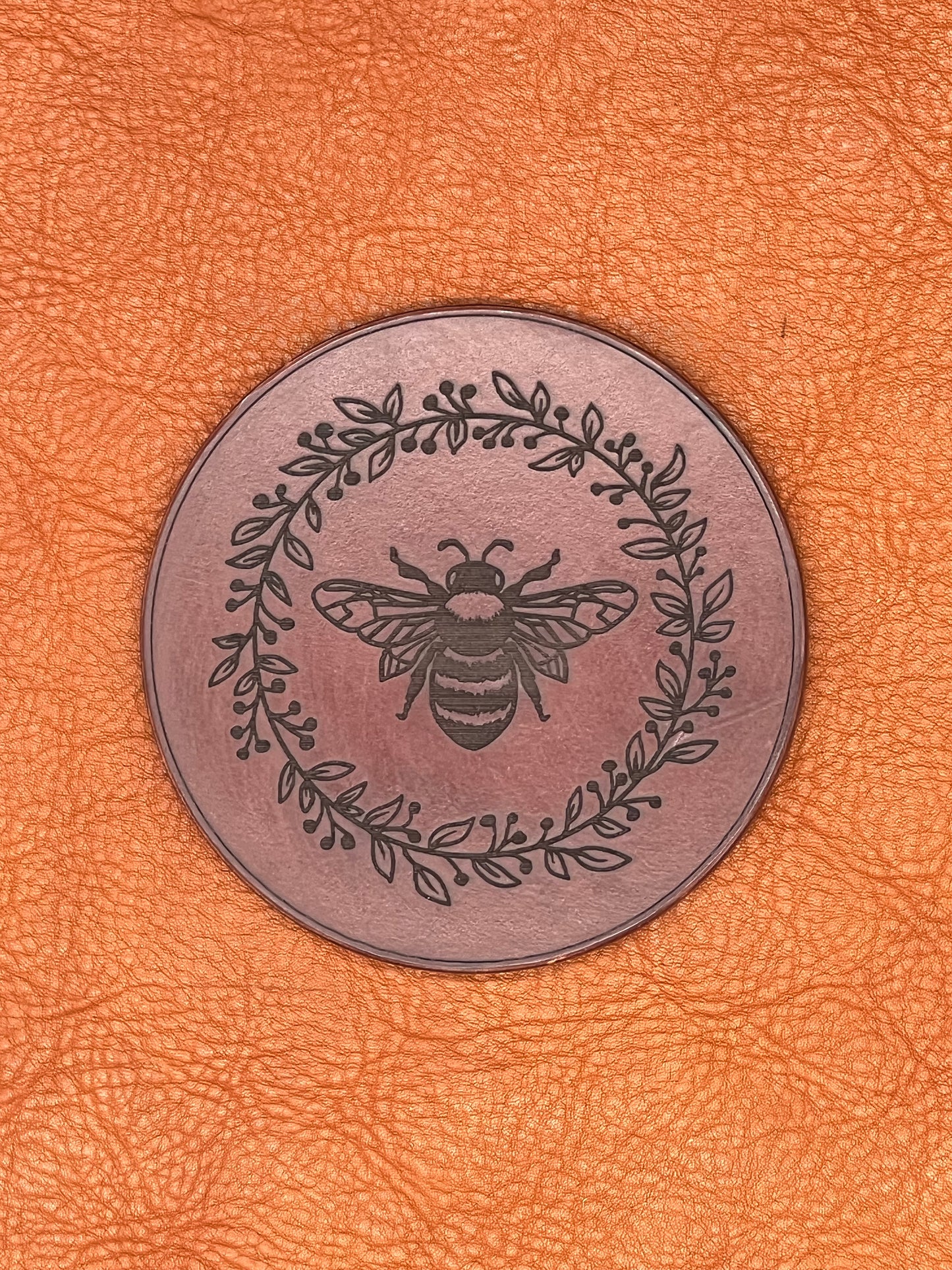 a chestnut colored leather coaster featuring an engraved bee encircled with vines and berries